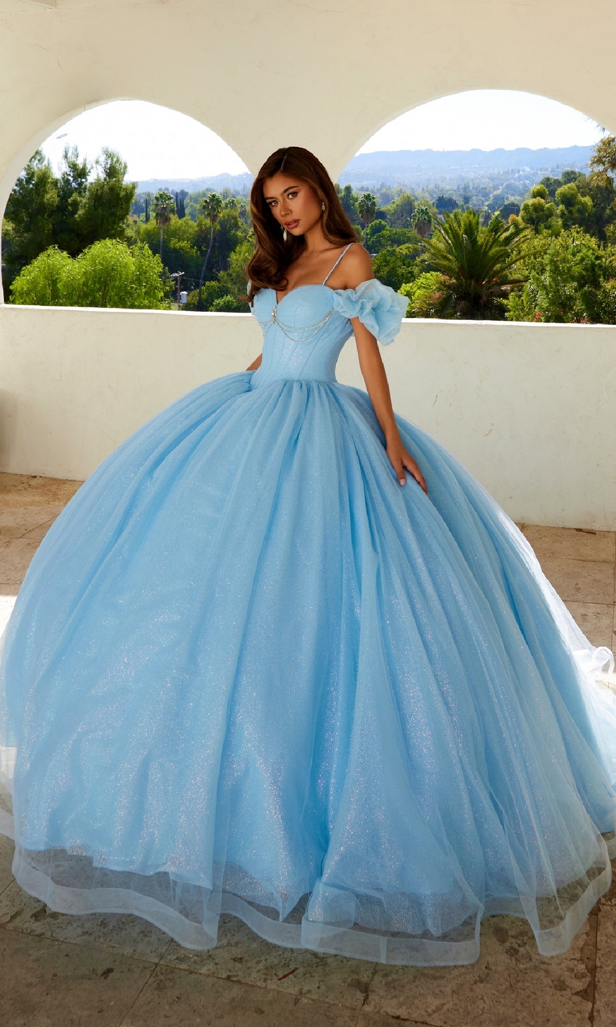 A young woman stands outdoors in the Quinceanera Dress JT1487H By Juliet, featuring a light blue princess style with puff off-the-shoulder sleeves and a full tulle skirt, set against lush greenery and an arched white wall.